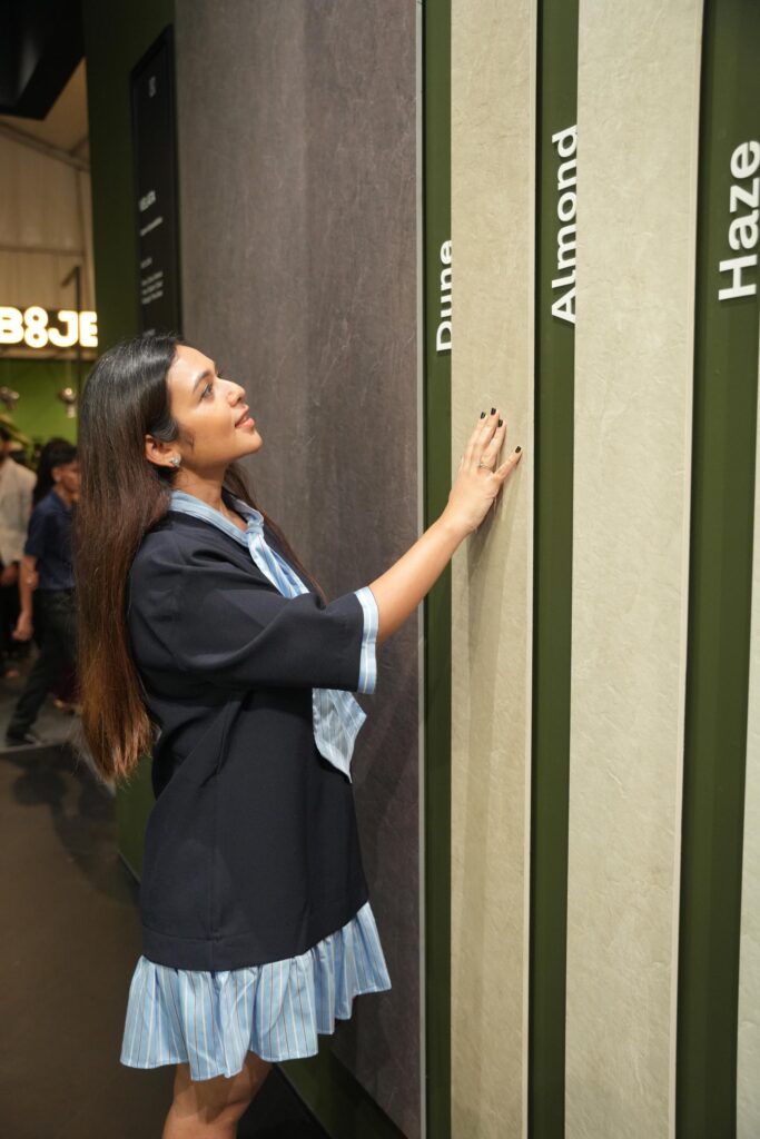 A visitor touching Velata sintered stone samples displayed vertically, with colour names Dune, Almond, and Haze visible on the display. 